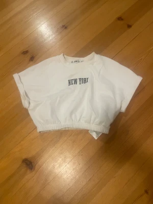 Vit croppad topp från FB Sister - Säljer en vit croppad topp från FB Sister i storlek XXS. Toppen har korta ärmar, elastisk nederkant och trycket 'NEW YORK' framtill. Perfekt för dig som gillar streetstyle och vill ha en enkel men snygg look.
