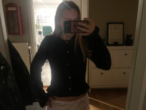 Svart fluffig kofta med pärlknappar - Mysig svart kofta med fluffig yta och pärlknappar framtill. Modellen är långärmad och har en lätt volangkant vid ärmslut och nederkant. Perfekt för dig som gillar en söt och trendig look. Passformen är normal och koftan är riktigt skön att ha på sig.