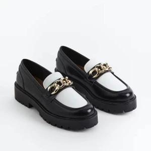 Chunky loafers med guldkedja - Säljer ett par svarta och vita chunky loafers med grov platt sula och rund tå. Skorna har en stor guldfärgad kedjedetalj framtill. 