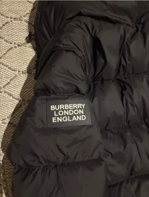 Svart Burberry pufferjacka med huva - Svart pufferjacka från Burberry med stor huva och klassiskt rutigt foder i grått, vitt och rött. Jackan har en tydlig Burberry London England-patch på ärmen, dragkedja framtill och praktiska fickor. Perfekt för kalla dagar och riktigt snygg streetstil. Priset kan diskuteras kan gå ner mycket!!! 