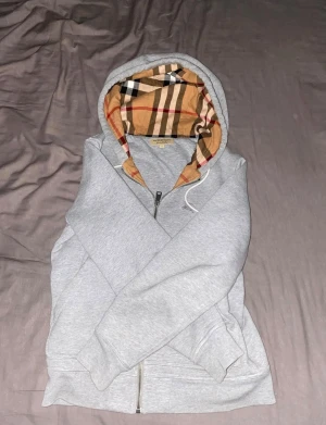Grå Burberry hoodie med dragkedja - Snygg grå hoodie från Burberry med klassiskt rutigt foder i huvan och insidan. Tröjan har dragkedja framtill, ribbade muddar och huva med det ikoniska Burberry-mönstret i beige, svart och rött. Materialet är mjuk bomull och polyester.