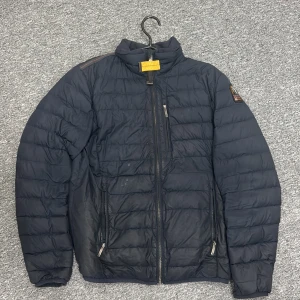 Mörkblå dunjacka från Parajumpers - Den har små hål i utsidan men den är inte märkbar annars är den ganska använd men i jätte bra form Snygg mörkblå dunjacka från Parajumpers med quiltad design och flera dragkedjor. Jackan har en patch på ärmen och en gul detalj vid kragen. Insidan är ljusgrå och jackan har hög krage och långa ärmar. Perfekt för kyliga dagar.