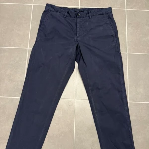 Mörkblå chinos från J.Lindeberg - Snygga mörkblå chinos från J.Lindeberg i bomullssatin. Klassisk rak passform med fickor fram och bak, bälteshällor och knappstängning. Perfekta för dig som gillar stilrena och bekväma byxor med lite dressad känsla. Storlek 31/32