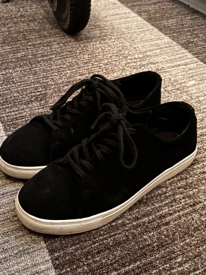 Svarta mockasneakers från Ciszere - Svarta sneakers från Ciszere med ovandel i mjuk mocka och klassisk vit platt sula. Skorna har rund tå och snörning framtill för en clean och stilren look. Perfekta för dig som gillar minimalistisk design och vill ha ett par schyssta sneakers till din vardagsstil.