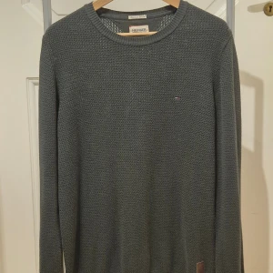Grå stickad tröja Tommy Hilfiger S (M/L) - Snygg grå stickad tröja från Tommy Hilfiger Denim med diskret logga på bröstet och en brun läderpatch nedtill. Tröjan har rund hals, ribbade muddar och långärmad passform. Perfekt för dig som gillar klassisk stil med en modern touch. Storlek S men sitter som en M/L.