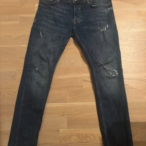 Blå jeans från SMOG med slitningar - Snygga blå jeans från SMOG i storlek 32/32. Jeansen har coola slitningar och detaljer på framsidan, klassisk femficksdesign och brunt läderpatch bak i midjan. Passformen är rak och materialet är robust denim. Perfekt för dig som gillar en avslappnad streetstil. Säljer då de var förstora vid köp. Hör av er vid minsta fundering😊