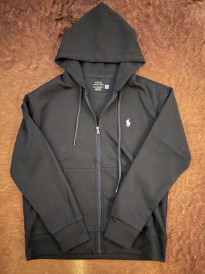 Svart hoodie från Polo Ralph Lauren - Svart hoodie från Polo Ralph Lauren med dragkedja och huva. Tröjan har snörning vid huvan, långärmade ärmar och klassisk Polo-logga broderad på bröstet. Tillverkad i mjuk bomull, perfekt för chill dagar. Snygg och enkel design som funkar till allt.