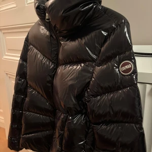 Svart pufferjacka från Colmar - Snygg svart pufferjacka från Colmar med glansig finish och hög krage. Jackan har en tydlig logga på ärmen och dragkedja framtill. Perfekt för kalla dagar när du vill hålla stilen och värmen.