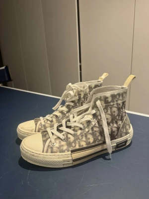 Dior B23 high-top sneakers, vit/grå - Säljer ett par Dior B23 high-top sneakers i storlek 43. Skorna har ett genomskinligt textilmaterial med det klassiska Dior-monogrammet i grått och vitt, snörning och robust vit gummisula med Dior-logga på sidan. Snygg och unik design med rund tå och platt sula. Låda samt skopåsar finns också. Tvättas såklart innan dem skickas. 