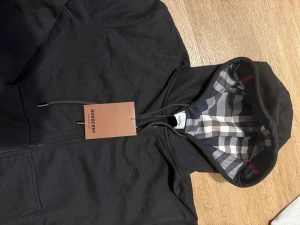 Svart Burberry hoodie med rutig huva - Säljer en svart hoodie från Burberry med klassisk rutig insida i huvan. Tröjan har dragkedja framtill, snörning vid huvan och två fickor. Materialet är mjuk bomull och passformen är normal. Perfekt för dig som vill ha en stilren och exklusiv look.