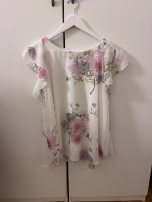Blommig vit blus. Storlek S - Säljer en söt och luftig blus i storlek S. Blusen är vit med blommigt mönster i rosa, lila och grönt. Den har volangärmar och en knapp i nacken. Materialet är tunt och flowy, perfekt för vår och sommar. Supersnygg till jeans eller kjol! Fått en liten bläck fläck (se bild ). Inget man tänker på☺️🥰