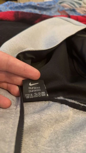 Grå nike tech från Nike XL - Snygg grå vindjacka från Nike i storlek XL med svart logga på bröstet. Jackan har huva, hel dragkedja framtill och två svarta fickor med dragkedja. Materialet är mjukt och känns som polyester, perfekt för blåsiga dagar.