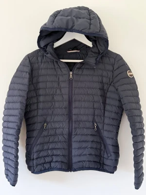 Mörkblå Colmar pufferjacka med huva - Snygg mörkblå pufferjacka från Colmar med huva och två fickor med dragkedja. Jackan har ett quiltat mönster och Colmar-logga på ärmen. Perfekt för kyliga dagar och har en stilren look som funkar till många outfits. Inlämnad på en kemtvätt innan jag la ut den så den är som ny 