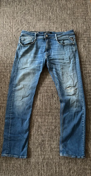 Replay Rocco blå jeans - Säljer ett par Replay Rocco jeans i blå tvätt med med hål på ena benet. Klassisk femficksmodell med raka ben och normal passform. Jeansen har snygga detaljer och är tillverkade i bomull. Perfekt för dig som gillar en avslappnad streetstil.