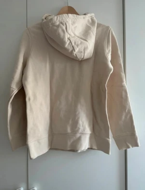 Beige Adidas hoodie med logga - Säljer en beige hoodie från Adidas med det klassiska trefoil-loggan i vitt framtill. Tröjan har huva med snörning och långa ärmar. Perfekt för både vardag och chill dagar