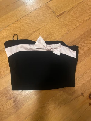 Svart tubtopp med rosett från SHEIN - Säljer en svart tubtopp från SHEIN i storlek XS. Toppen har en ljus silkesliknande detalj upptill och en snygg rosett i mitten. Perfekt för dig som gillar stilrena och söta detaljer. Materialet känns mjukt och har en lätt glans.