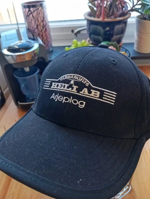 Svart keps Vildmarksflyg Heli AB Arjeplog - Svart keps med böjd skärm och broderad vit text fram och bak. Märkt med 'Vildmarksflyg Heli AB Arjeplog' framtill och 'Vildmarksflyg' baktill. Justerbar baktill för perfekt passform. Snygg och enkel design, perfekt för dig som gillar äventyr.
