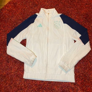 Vit Ajax vindjacka  - Snygg vit vindjacka från Adidas i storlek XS med marinblå detaljer på axlarna och mintgröna ränder på ärmarna. Jackan har dragkedja framtill, elastiska muddar och en cool logga på bröstet. Perfekt för dig som gillar sportig stil och streetwear.