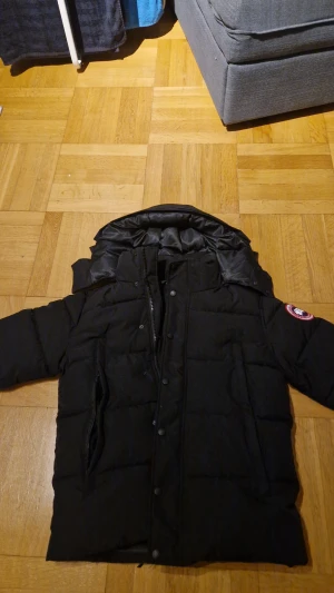 Svart pufferjacka från Canada Goose - Svart pufferjacka från Canada Goose i nyskick, ikonisk röd och vit patch på ärmen. Storlek XS men passar bättre S. Jackan har huva, dragkedja och knappar framtill, Perfekt för kalla vinterdagar.