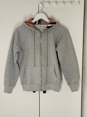 Grå hoodie från Burberry med dragkedja - Snygg grå hoodie från Burberry med klassiskt rutigt mönster i huvan. Tröjan har hel dragkedja, snörning vid huvan och två fickor framtill. Perfekt för dig som gillar stilrena och exklusiva märken. Materialet är mjuk bomull och polyester. Tveka inte på att skicka ett prisförslag.