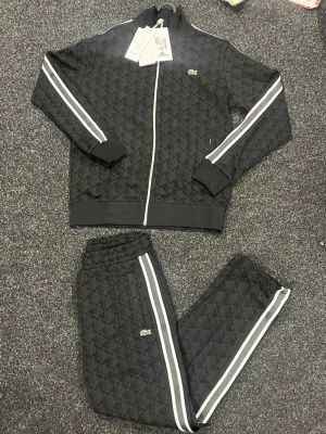 Svart mönstrad tracksuit från Lacoste - Snygg svart tracksuit från Lacoste med geometriskt mönster och vita ränder längs ärmar och ben. Setet har dragkedja på jackan och byxorna har elastisk midja. Materialet är mjukt och lätt, perfekt för chill eller sport. Klassisk Lacoste-logga på både jacka och byxa.     Alla storlekar finns 