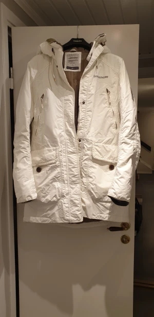 Vit parkajacka från Didriksons - Säljer en snygg vit parkajacka från Didriksons med huva och flera praktiska fickor med både dragkedja och knappar. Jackan har lång ärm och är tillverkad i slitstark polyester, perfekt för kyliga dagar. Stilsäker look med diskret logga på bröstet.