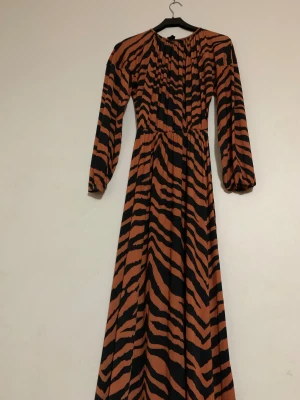 Zebramönstrad långklänning med puffärm - Säljer en maxilång klänning med zebramönster i svart och brun. Klänningen har rundad hals, långa puffärmar och markerad midja. Materialet känns mjukt och flowy, perfekt för dig som gillar att sticka ut med coola mönster och färger.