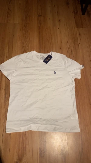 Vit t-shirt från Polo Ralph Lauren - Vit t-shirt från Polo Ralph Lauren i bomull med klassisk blå broderad logga på bröstet. Modellen är slim fit med korta ärmar och rund hals. Perfekt basic-plagg med stilren design och skön passform.