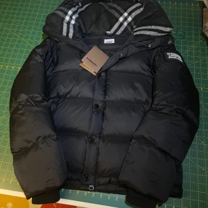 Burberry jacka/väst - Exklusiv Burberry dunjacka i helt oanvänt nyskick, tags kvar. Ikoniskt rutmönster i huvan och avtagbara ärmar så den kan bäras som väst. Varm, lyxig och perfekt för både vinter och streetwear.  Pris kan diskuteras. Hör av dig om du vill se fler bilder!