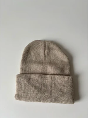 Beige stickad mössa - En stilren beige mössa med klassisk uppvikt kant. Stickad i mjukt material som håller dig varm och passar perfekt till alla dina outfits. Enkel design utan detaljer eller mönster, vilket gör den lätt att matcha. Helt ny.