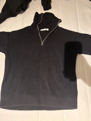 Svart ribbad stickad tröja Weekday - Svart stickad tröja från Weekday med ribbad struktur och half zip-dragkedja i halsen. Tröjan har lång ärm och hög krage som ger en cool och avslappnad vibe. Perfekt för dig som gillar stilrena och enkla plagg med en twist.
