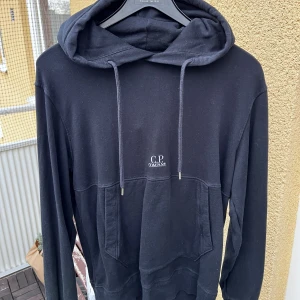 Svart hoodie från C.P. Company - Svart hoodie från C.P. Company med broderad logga på bröstet och patch med logga i nacken. Tröjan har huva med snörning, stor magficka och långa ärmar. Tillverkad i mjuk bomull, perfekt för chill dagar. Snygg och enkel design som funkar till det mesta.Priset är ristat i sten går ej att pruta!!