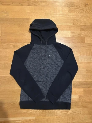 Mörkgrå hoodie från Hollister - Snygg mörkgrå och svart hoodie från Hollister i storlek S. Tröjan har huva med snörning, långärmade svarta ärmar och ett litet Hollister-logo på bröstet. Materialet är mjukt och skönt, perfekt för chill dagar. Passformen är normal och designen är stilren.