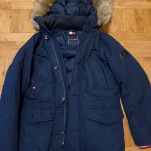 Blå vinter jacka Tommy Hilfiger  - Snygg marinblå parkajacka från Tommy Hilfiger med fuskpäls på huvan, quiltad insida och coola detaljer i rött och vitt. Jackan har flera fickor, dragkedja och knappar framtill samt ett stort Tommy Hilfiger-märke på ärmen. Perfekt för kalla dagar. Strlk XS men passar mig som är 185cm