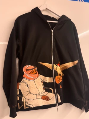 Svart hoodie med broderad motiv - Unik svart hoodie med dragkedja och huva, prydd med ett stort broderat motiv av en man i traditionell klädsel och en fågel på framsidan. Kontrastsömmar och detaljer i beige, orange och rött. Perfekt för dig som gillar statement-plagg och coola broderier.