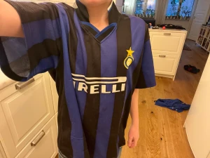 Inter Ronaldo 9 fotbollströja blå/svart - Klassisk Inter fotbollströja med Ronaldo 9 på ryggen. Tröjan är blå och svart med breda ränder, Pirelli-logga på bröstet och klubbmärke med stjärna. Tillverkad i lätt och ventilerande material, kortärmad och med unik krage. Perfekt för dig som älskar retro och fotbollshistoria.