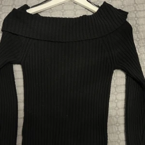 Svart ribbad offshoulder stickad tröja - Snygg svart stickad tröja med ribbad struktur och offshoulder-design. Tröjan har långa ärmar och en bred krage som ger en cool och trendig look. Perfekt för dig som gillar stilrena och bekväma plagg med lite edge.