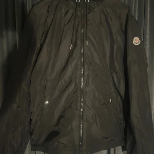 Moncler Grimpeurs 3/L - Svart vindjacka från Moncler med klassisk logotyp på ärmen och snygga detaljer som dragsko i huvan och dragkedja framtill. Jackan har två fickor med tryckknappar och är tillverkad i lätt polyester som passar perfekt för blåsiga dagar.