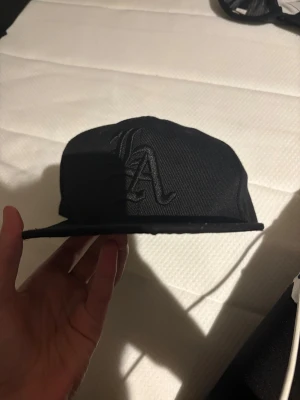 Svart LA keps med broderad logga - Säljer en svart LA keps från New Era med broderad LA-logga framtill i samma svarta färg. Klassisk flat brim och justerbar baksida. Kepsen är tillverkad i bomull och polyester, perfekt för en streetwear-look.