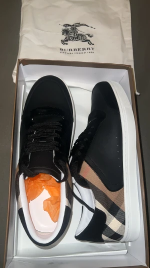 Burberry rutig sneakers svart/vit - Snygga Burberry sneakers med klassiskt rutigt mönster i beige, svart och vitt. Skorna har svart ovandel i skinn och textil, vita platta sulor och svarta snören. Perfekt för dig som vill ha en stilren och lyxig look med ikonisk Burberry-touch.