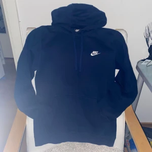 Mörkblå Nike hoodie med huva - Snygg mörkblå hoodie från Nike i storlek S. Tröjan har huva med snörning, en stor magficka och Nike-logga broderad i vitt på bröstet. Tillverkad i mjuk bomull och polyesterblandning, perfekt för chill eller träning. 