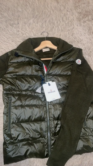 Moncler cardigan  - Snygg grön pufferjacka från Moncler med stickade ärmar och krage. Jackan har dragkedja framtill med röd, vit och blå detalj, samt Moncler-logga på ärmen. Materialmix av polyester och stickad ull ger en modern look. Perfekt för kyliga dagar.