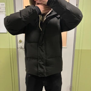 Canada Goose wyndham - Säljer en svart wyndham Canada Goose med huva och klassisk röd logga på ärmen. Jackan har en loose passform, knappar framtill och är långärmad. Perfekt för kalla dagar med sin tjocka och quiltade design.