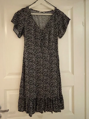 Svart blommig kortklänning med volang - Söt svart kortklänning med små blommor och volang längst ner. Klänningen har v-ringning och korta ärmar. 