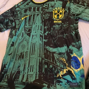 Nike Brasil CBF Dri-FIT fotbollströja - Unik Brasilien CBF fotbollströja från Nike med Dri-FIT-material. Tröjan har korta ärmar, svart och grön bas med gula detaljer och ett grafiskt tryck av brasilianska landmärken samt flaggan. Rund hals med svart och gul kant. Perfekt för dig som vill sticka ut på planen.
