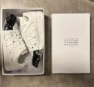 Maison Margiela black paint splatter - Maison Margiela sneakers i vitt skinn med svart färgstänk över tå. Väldigt bra skor med hög kvalité som är helt nya och oanvända. Kommer med originalkartong. Hör av dig vid frågor eller funderingar!