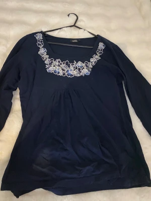 Mörkblå blus med broderade blommor - Säljer en mörkblå blus från Isolde med lång ärm och rundad halsringning. Blusen har vackra broderade blommor i vitt och blått runt halsen som ger en romantisk vibe. Materialet känns mjukt och lätt, perfekt för dig som gillar detaljer.