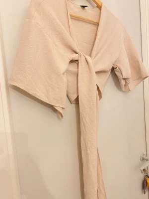 Beige omlott bolerotopp med knyt - Säljer en beige bolerotopp med omlott-design och knyt framtill. Toppen har korta, vida ärmar och är tillverkad i ett strukturerat, lätt material som ger en cool och avslappnad vibe. Perfekt att styla över linne eller klänning för en trendig look.