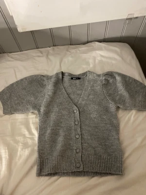 Grå stickad kofta från Gina Tricot - Superfin grå stickad kofta från Gina Tricot i storlek XS. Koftan har korta puffärmar, v-ringning och knappar framtill i samma färg som plagget. Ribbad kant vid ärmslut och nederkant ger en snygg detalj. Perfekt för lager-på-lager under kyliga dagar.