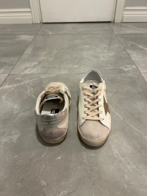 Golden Goose vita sneakers med glitterstjärna - Golden Goose sneakers i vitt skinn med beige mockadetaljer och silverfärgad häl. Sidan har en stor glittrig guldstjärna och snörning framtill. Platt sula och rund tå ger en klassisk look. Perfekt för dig som gillar exklusiva och trendiga sneakers.